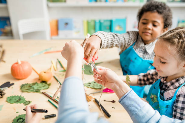 istock Cute schoolboy cutting thread with scissors while helping his teacher at lesson 1186533669