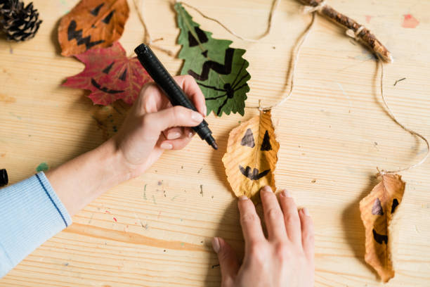 istock Hands of female with black highlighter drawing faces on dry leaves with threads 1186533715