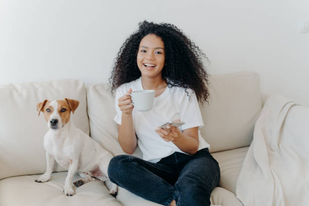 istock Delighted dark skinned woman poses in modern apartment, sits on comfortable sofa with pet, drinks coffee, uses mobile phone for online communication, being in good mood, flipping news, uses app 1186556418