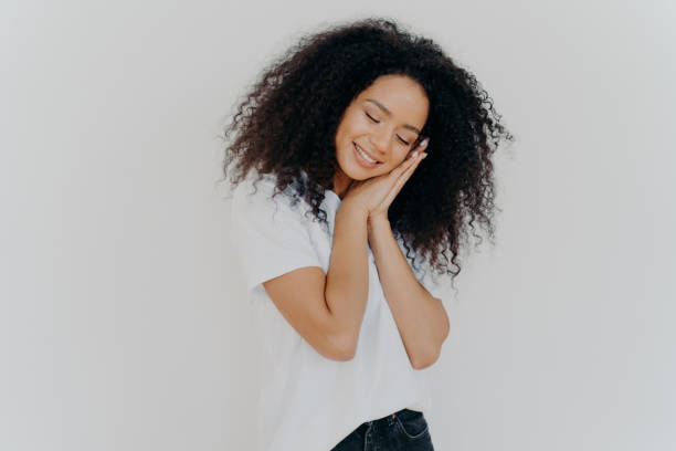 istock Charming beautiful woman with tender smile, leans on pressed palms, dreams about sleep, tilts head, wears white t shirt, has natural beauty, perfect teeth, poses indoor. Young dreamy Afro woman 1186556652