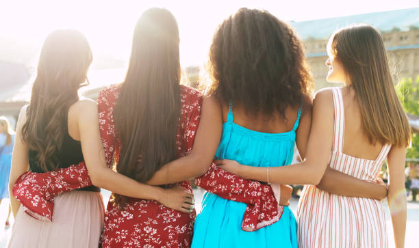 Sunny women days. A group of four best friends in magnificent summer outfits are hugging each other and posing with their backs to the camera. arm in arm stock pictures, royalty-free photos & images