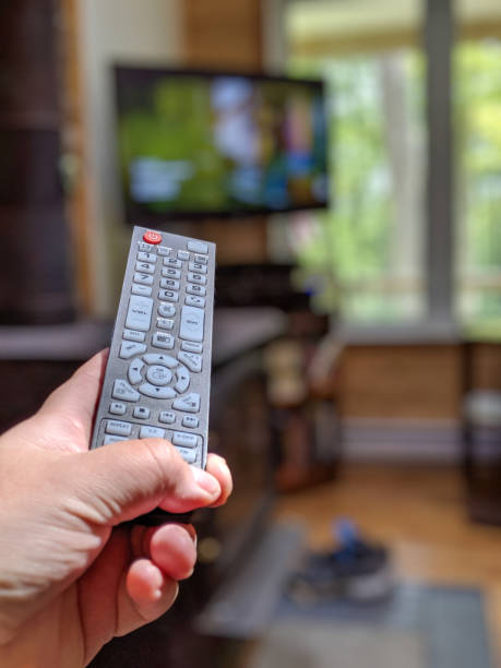 Hand points remote control towards TV in natural light living room setting.