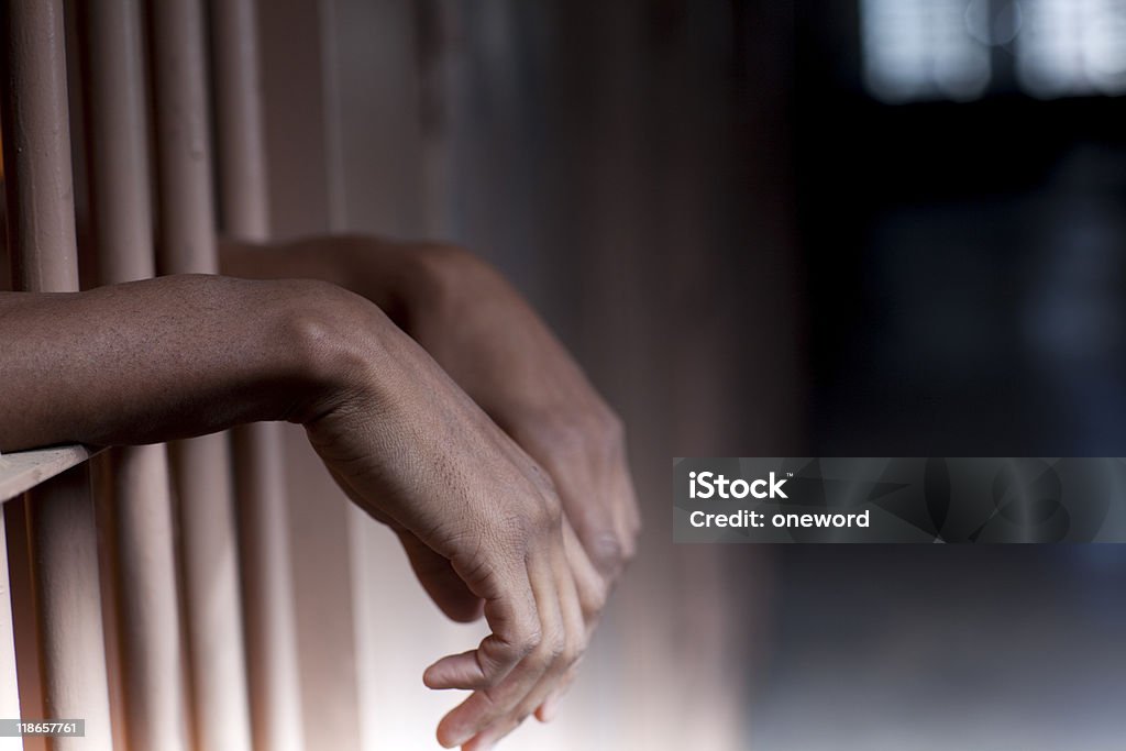 Hands resting on prison bars in a forlorn pose An African American male with hands hanging out prison bars Prison Stock Photo