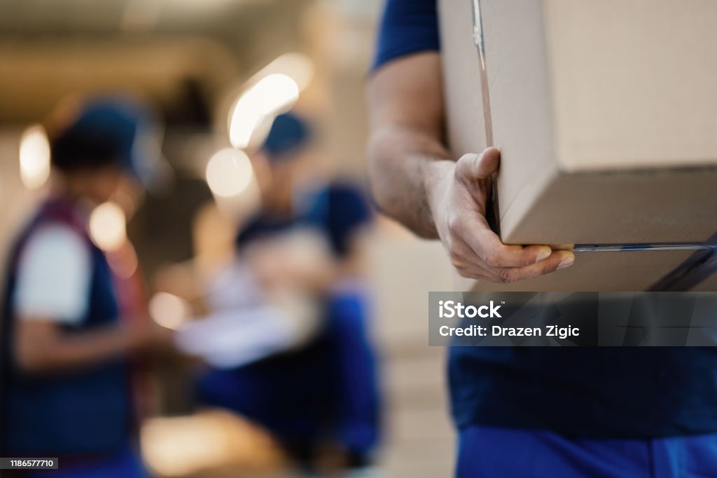 Close-up of unrecognizable delivery man with cardboard box. Close-up of a worker carrying cardboard box while making a delivery. There are people in the background. Box - Container Stock Photo