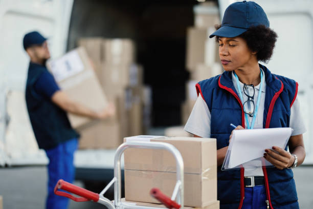 African American female worker writing data in a delivery list. Black female delivery woman taking notes in a clipboard while her coworker is unloading packages form a van. mover delivery person box hand truck stock pictures, royalty-free photos & images