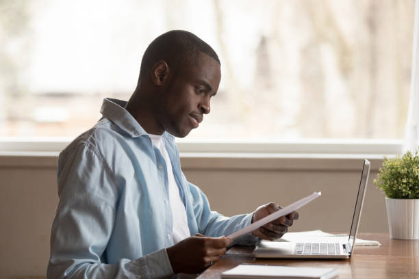 Focused biracial man reading paper letter working at laptop Focused african American millennial man sit at table work study using laptop reading paper document, concentrated biracial male look through consider paperwork letter, good news notice at workplace college acceptance letter stock pictures, royalty-free photos & images