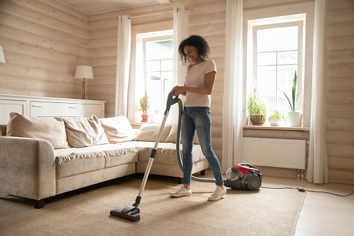 Happy african American young woman preform household chores cleaning living room using modern vacuum cleaner, smiling biracial wife hoover remove dust from home carpet, do housekeeping job
