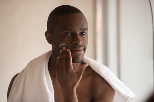 Young african American male look in mirror in bathroom apply face cream on cheeks, millennial biracial man shave with balm do skincare routine after having shower in home bath, hygiene concept