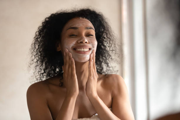 Smiling biracial woman apply face mask after shower Close up of smiling mixed race ethnicity young woman look in mirror apply facial moisturizing mask, positive african American female massage face do skincare beauty anti-aging treatment in bathroom facial cleanser stock pictures, royalty-free photos & images