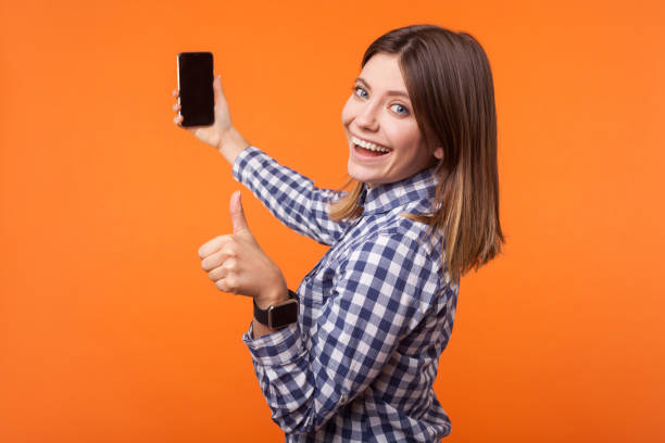 half-turned portrait of brunette woman with charming smile holding phone. isolated on orange background - half smile imagens e fotografias de stock