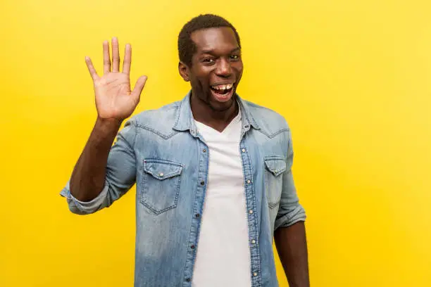 Hello! Portrait of positive handsome man in denim casual shirt with rolled up sleeves smiling friendly and waving hand saying hi, welcoming gesture. indoor studio shot isolated on yellow background