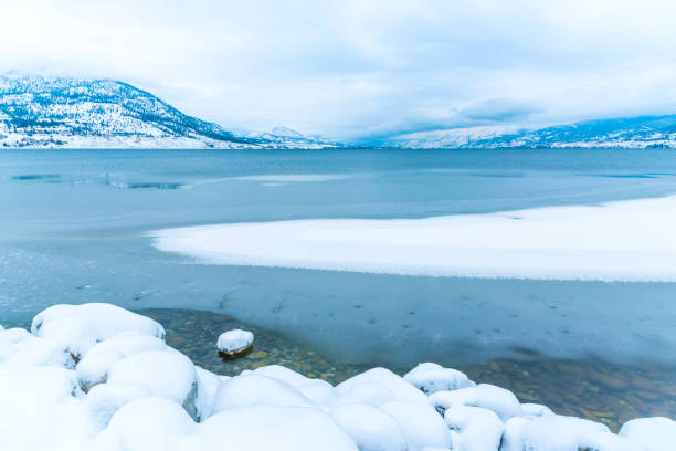눈으로 덮인 바위 해안선과 얼어붙은 호수의 전경, 겨울에는 눈으로 덮인 산과 안개를 볼 수 있습니다. - okanagan penticton lake okanagan lake 뉴스 사진 이미지