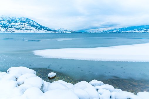 Ice and snow on Okanagan Lake in winter after a snowstorm with cloudy sky and mountains in distance, in Penticton, British Columbia, Canada