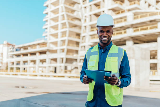 ingénieurs masculins portant un casque de protection et des vêtements réfléchissants, debout sur le chantier de construction devant un grand bâtiment. - architecture vibrant color bright built structure photos et images de collection