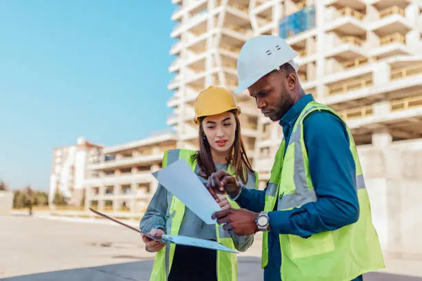 Two engineers, male and female, in protective helmets and reflective clothing, standing on construction site infront of a large building. Engineers looking at blueprints, planning and discussing project development.