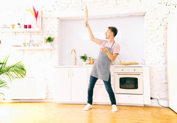 彼の手にプレートとローリングピンを持つ台所で幸せな若いブロンドの男は、楽しんで踊ります。クッキング・ラブ - pin head ストックフォトと画像