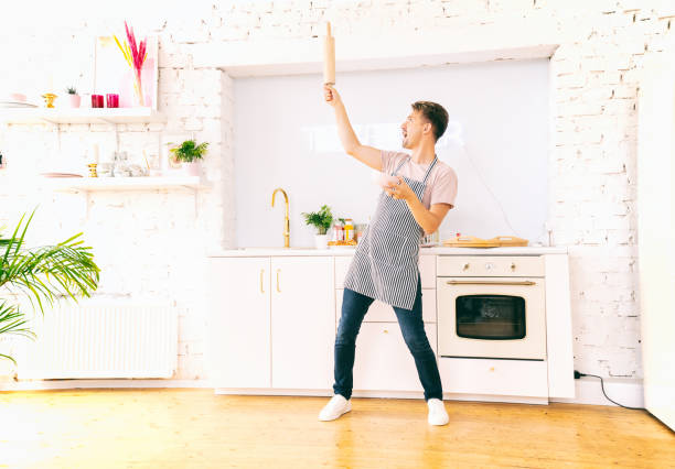 Happy young blond man in an apron in the kitchen with a plate and rolling pin in his hands, dancing having fun. Cooking Love