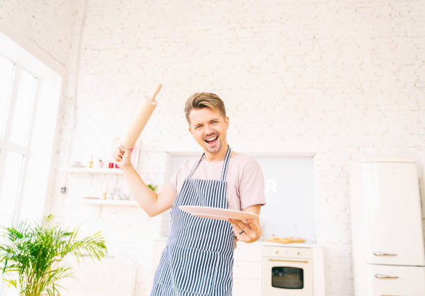 Happy young blond man in an apron in the kitchen with a plate and rolling pin in his hands, dancing having fun. Cooking Love