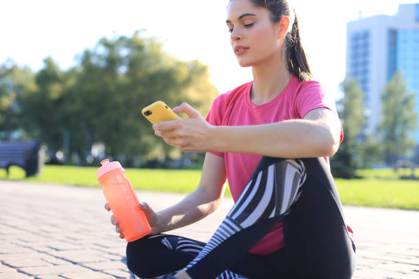 istock Beautiful young woman looking away while sitting on exercise mat at park, relaxing after training and using smartphone. 1186690608