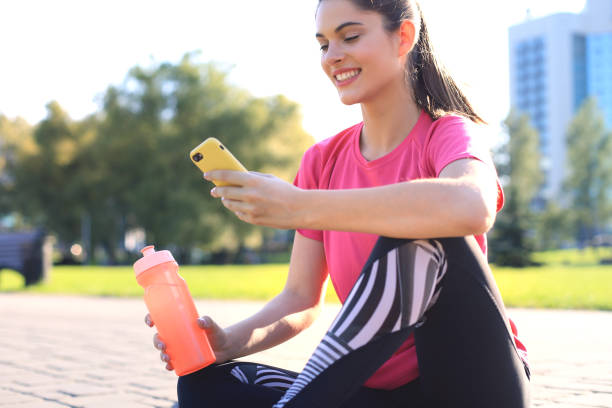 istock Beautiful young woman looking away while sitting on exercise mat at park, relaxing after training and using smartphone. 1186692982