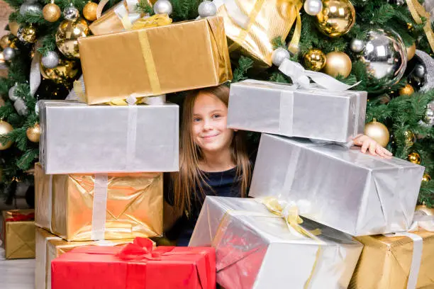 Photo of Happy preteen girl sitting among wrapped presents beside decorated Christmas tree on boxing day