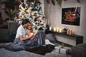 Couple in love drinking mulled wine or eggnog while sitting near fireplace. Beautiful woman and man covered with gray blanket and present gift. Christmas tree on background . Hygge concept