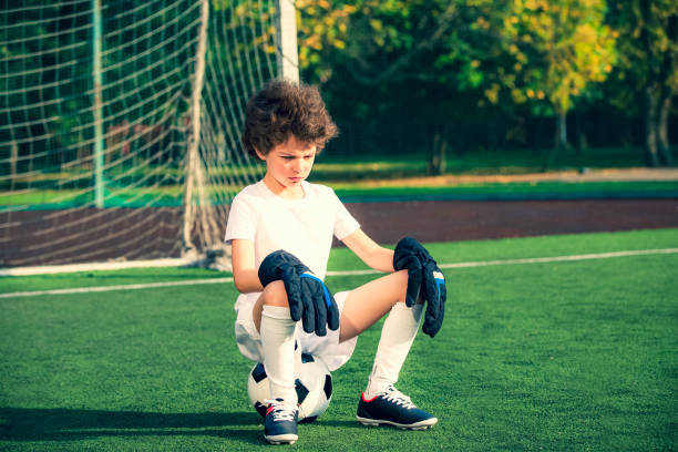 Summer soccer tournament.emotions and joy of the game. Young goals sitting on a ball. Boy goalkeeper in football sportswear on stadium with ball. Sport concept. Summer soccer tournament.emotions and joy of the game. Young goals sitting on a ball. Boy goalkeeper in football sportswear on stadium with ball. Sport concept. offense sporting position stock pictures, royalty-free photos & images