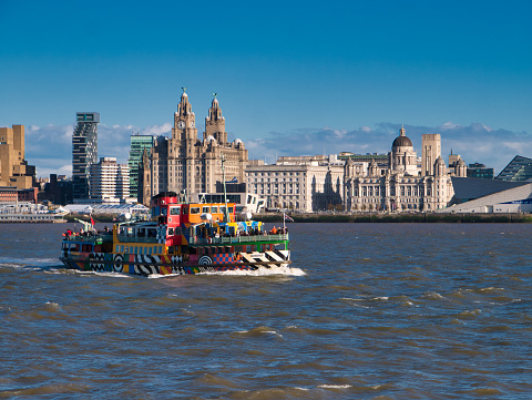 The River Mersey Ferry Snowdrop passes the Three Graces on the historic Liverpool waterfront on a sunny afternoon. The ferry is decorated in the style of a dazzle ship with a distinctive pattern entitled \