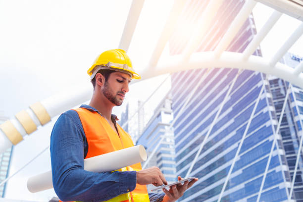 Blue collar worker, engineer man work on digital tablet, hold blue print, wear safety hat, stand near worksite. Worker guy survey a land for big project of building or estate. look determined