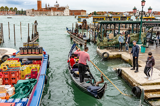 This is an image of the Gondola stations at San Marco Square in Venice, Italy with a boarding station and tourists in the gondolas and taking photos on the canal side. On the left is a delivery barge.