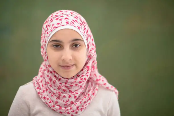A young female Muslim elementary student stands in front of a black board for a head shot portrait.  She is dressed casually and wearing a head scarf.