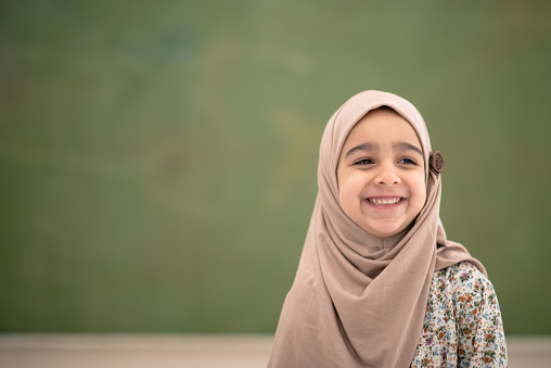 A young female Muslim elementary student stands in front of a black board for a head shot portrait.  She is dressed casually and wearing a head scarf.