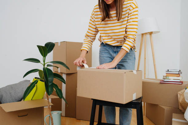 Cropped image of a woman packing, she's moving out from old apartment. Middle age woman in casual clothes standing next to pile of boxes, packing, she's moving out from an old apartment. Sealing cardboard boxes with adhesive scotch tape. Close up, cropped, no head. packing stock pictures, royalty-free photos & images