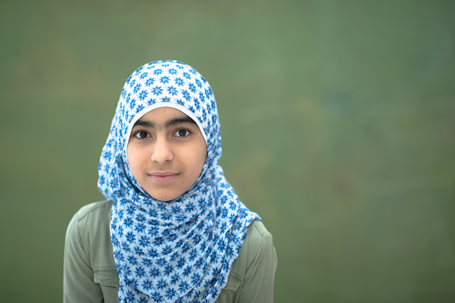 A young female Muslim elementary student stands in front of a black board for a head shot portrait.  She is dressed casually and wearing a head scarf.
