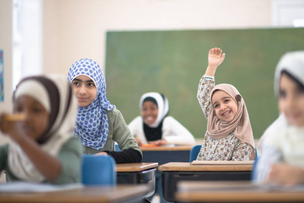 A young Muslim elementary student sits at her desk with her hand raised to ask the teacher a question.  She is smiling, wearing casual clothing and a head scarf.