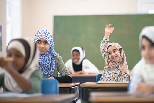 A young Muslim elementary student sits at her desk with her hand raised to ask the teacher a question.  She is smiling, wearing casual clothing and a head scarf.