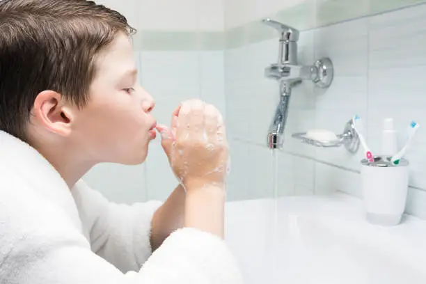 Photo of a boy in a white bathrobe in the bathroom tries to inflate a soap bubble with soaped hands