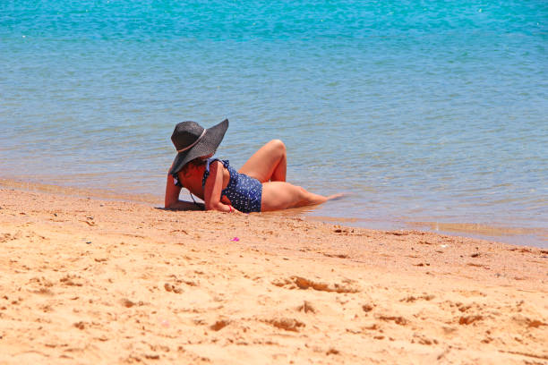 Hurghada / Egypt. 28 July 2018: Modern woman in dark hat lying by sea on beach. Woman enjoying summer holidays. Lonely woman lying on beach and relaxing. Enjoying summer vacations