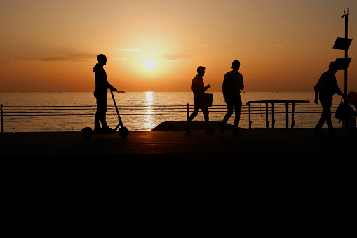 The silhouettes of group of people riding scooter and walking with stroller on the sea shore against the sunset. The sunset over sea is visiable on the background.