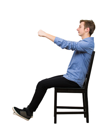 Side view full length portrait of confident teenager boy dreamer pretend to drive an imaginary transportation, car or bike, while sitting in his chair isolated over white background.