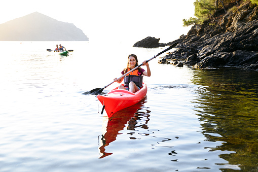 Close-up of smiling young Spanish female kayaker paddling in Mediterranean waters in early morning with friends in background.