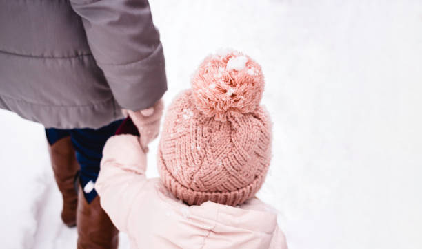 dziewczynka ubrana w kurtkę puffer i czapkę z dzianiny idzie w parze z matką lub babcią. zima i śnieg zimna pogoda - hat toddler little girls pink zdjęcia i obrazy z banku zdjęć