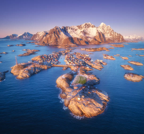 vista aérea de rocas en el mar, montañas nevadas, cielo púrpura al atardecer en las islas lofoten, noruega. paisaje de invierno con pequeñas islas en el agua, campo de fútbol en la roca. vista superior de la ciudad de henningsvaer - lofoten henningsvaer norway village fotografías e imágenes de stock