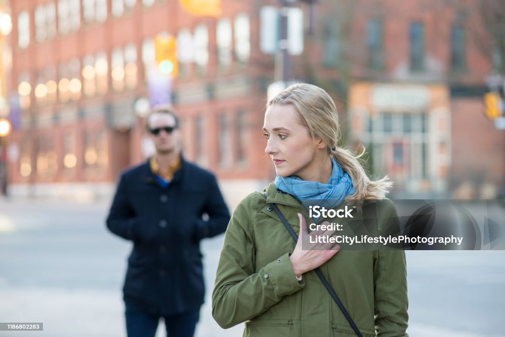 Feeling Unsafe When Walking Scared young woman with blonde hair looking back over her shoulder at a stranger in a black trench coat that is following behind her downtown in an urban city. Stalker - Person Stock Photo