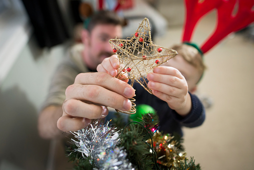 Young boy and his father putting the star on a Christmas tree as they are decorating for the holidays. They are both wearing festive reindeer antlers.