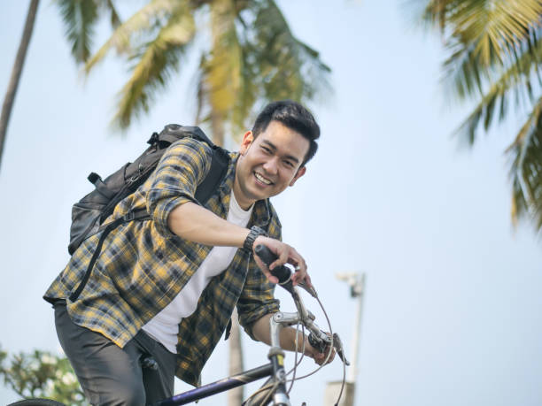 緑の木で自転車に乗ってバックパックを持つ幸せなアジアの男 - campus university walking school ストックフォトと画像