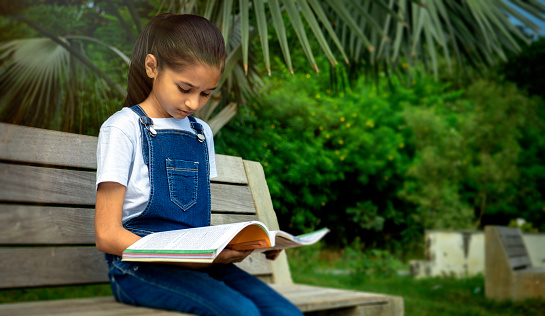 The Indian little girl sitting on a bench in the park and reading books.Reading the book in outdoors