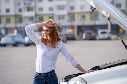 Young stressed woman driver standing near broked car with open hood.