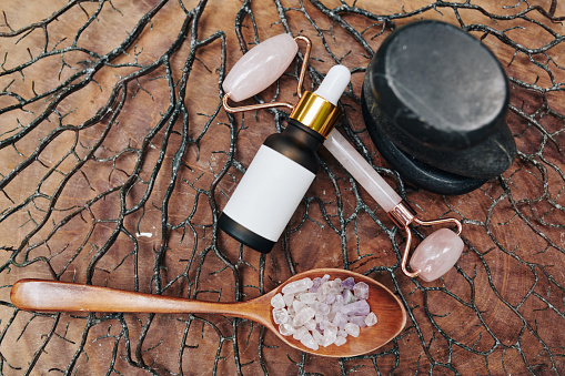 Pipette bottle with rejuvenating serum, rose quarts massage roller and spoon of gemstones on wooden surface with small branches