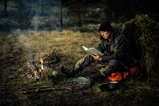 Man reading book while sitting near campfire in forest.